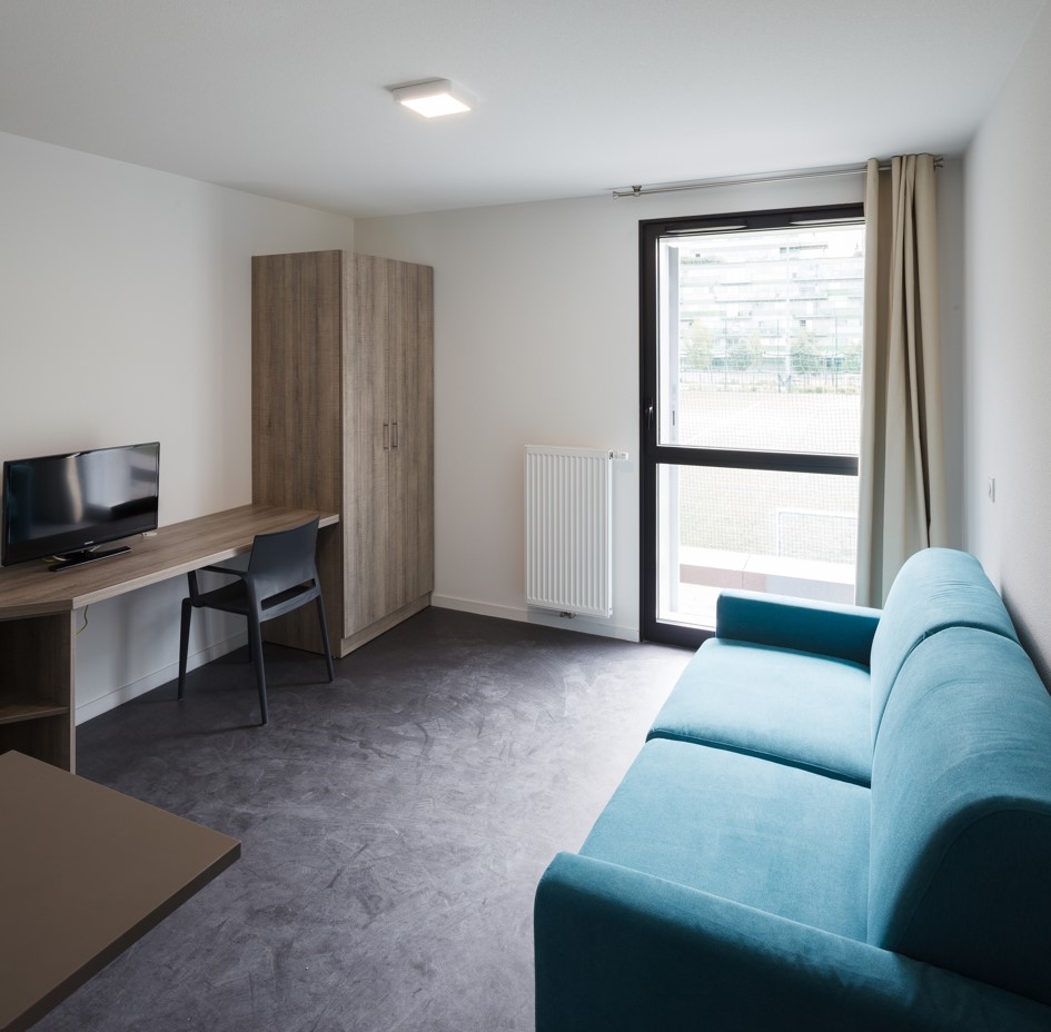 Bright student studio with turquoise sofa, fitted desk, wooden wardrobe, window and glass door to outside.