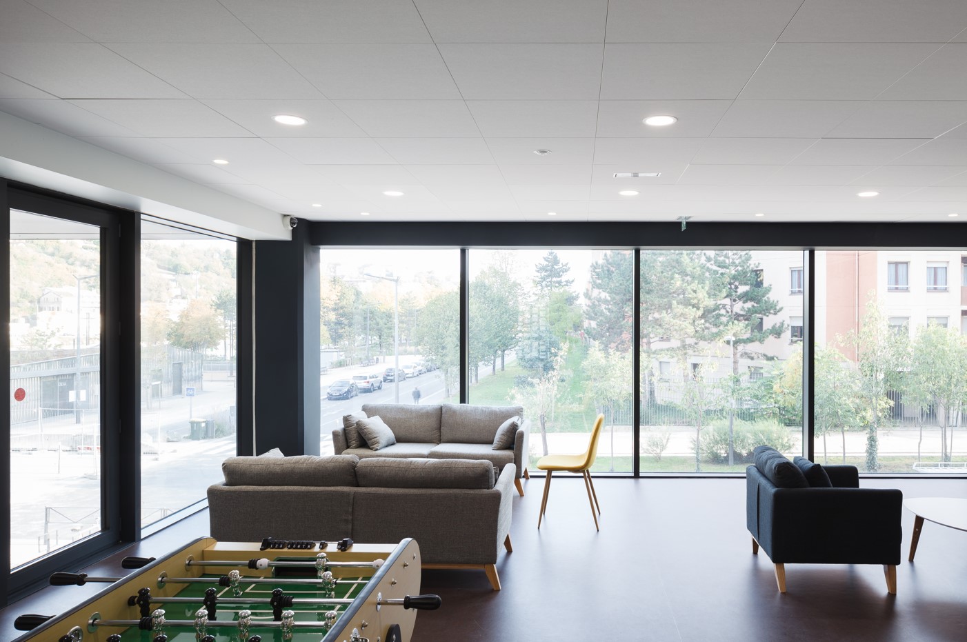 Bright communal area with large picture windows, grey sofas, armchairs, a yellow chair and table football in the foreground.