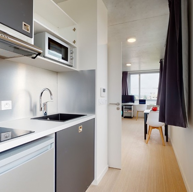Interior of a student studio with fully-equipped kitchenette, hob, sink, microwave, and view of a bright study area in the background.