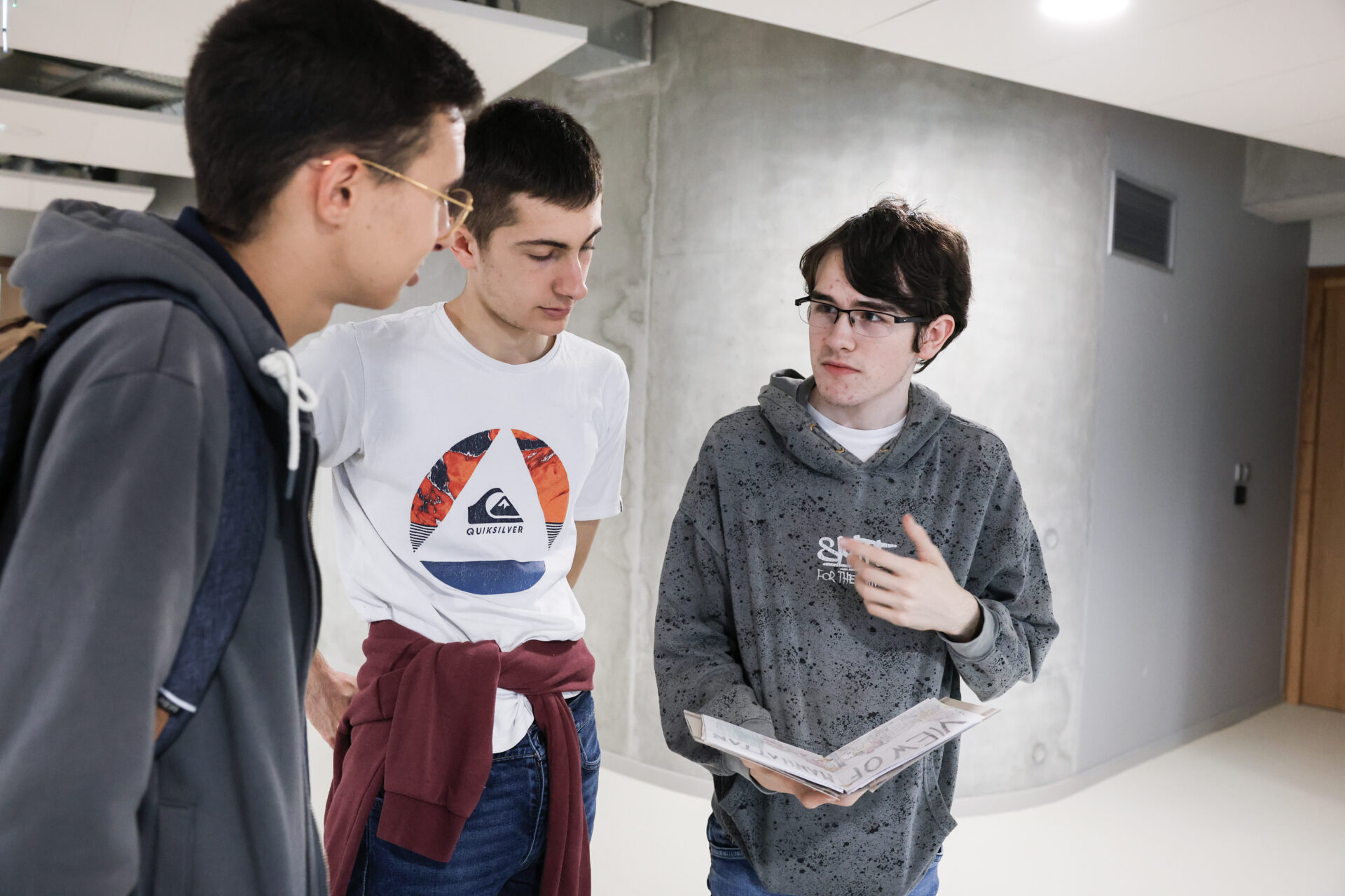 Three students talk in a corridor while holding a sketchbook or an art project, in a school context.