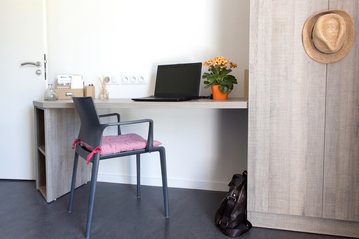 Bright student desk with chair, closed laptop, green plant, supplies and light wood cupboard.