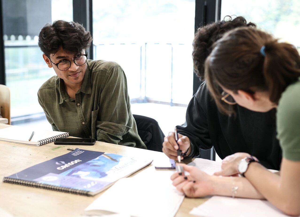 Trois étudiants travaillent ensemble autour d’une table, échangeant des idées avec des carnets de croquis et du matériel de dessin, dans un environnement lumineux.