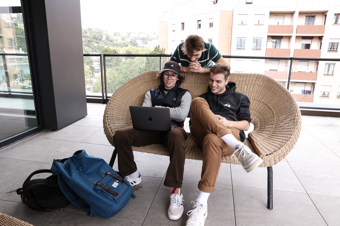 Three students sitting on a rattan bench on a terrace, looking at a laptop together in a relaxed atmosphere.