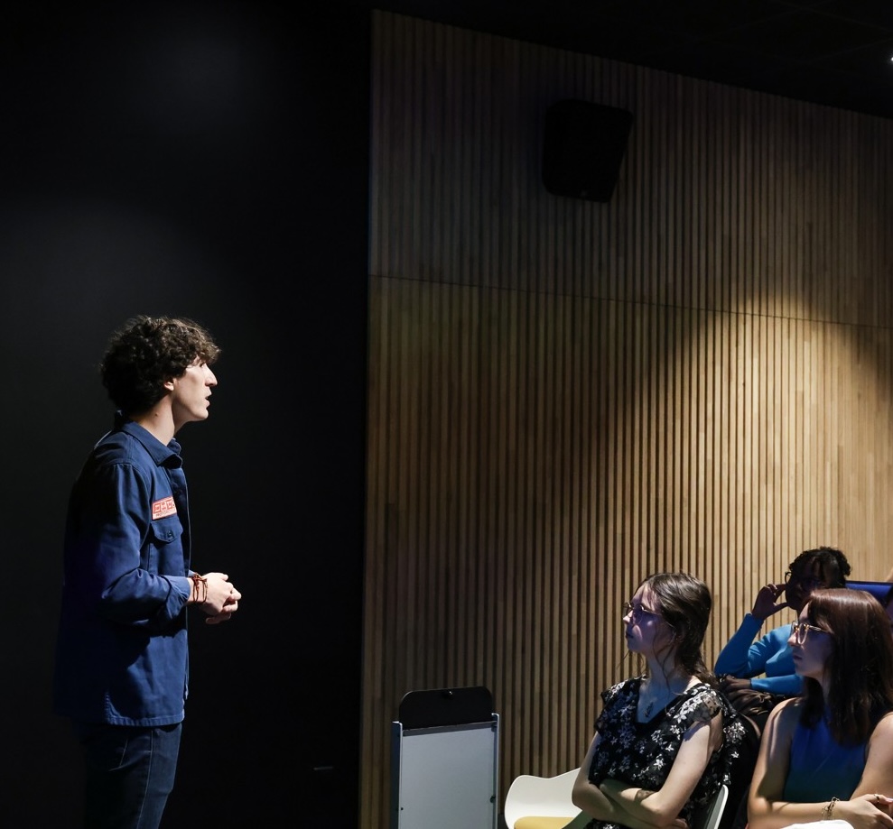 A student presents standing in front of an attentive audience in a conference room with wood-panelled walls and subdued lighting.