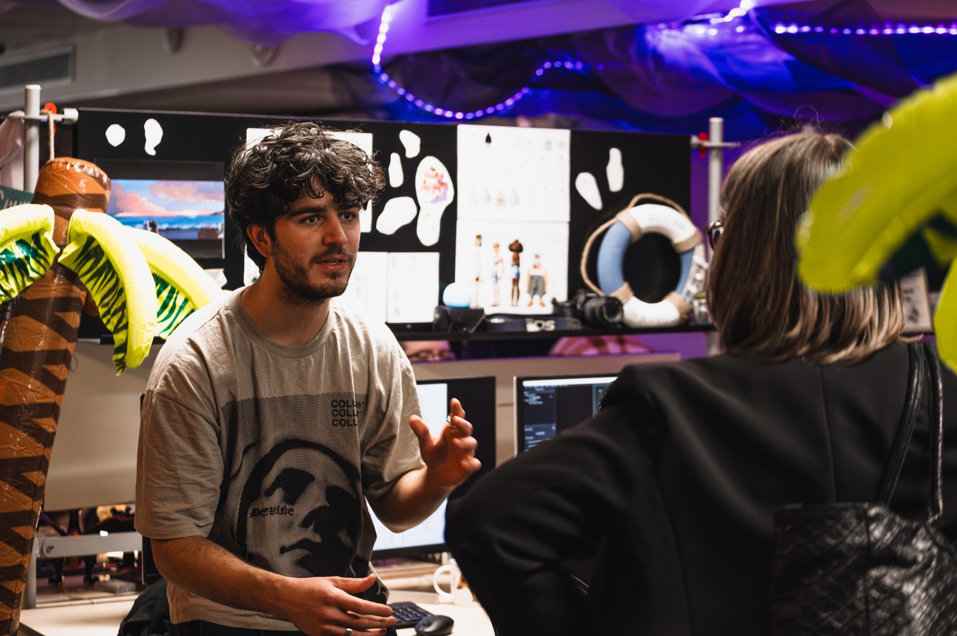 A 3D animation student presents his project to a visitor, in a space decorated with inflatable palm trees and visual research boards.
