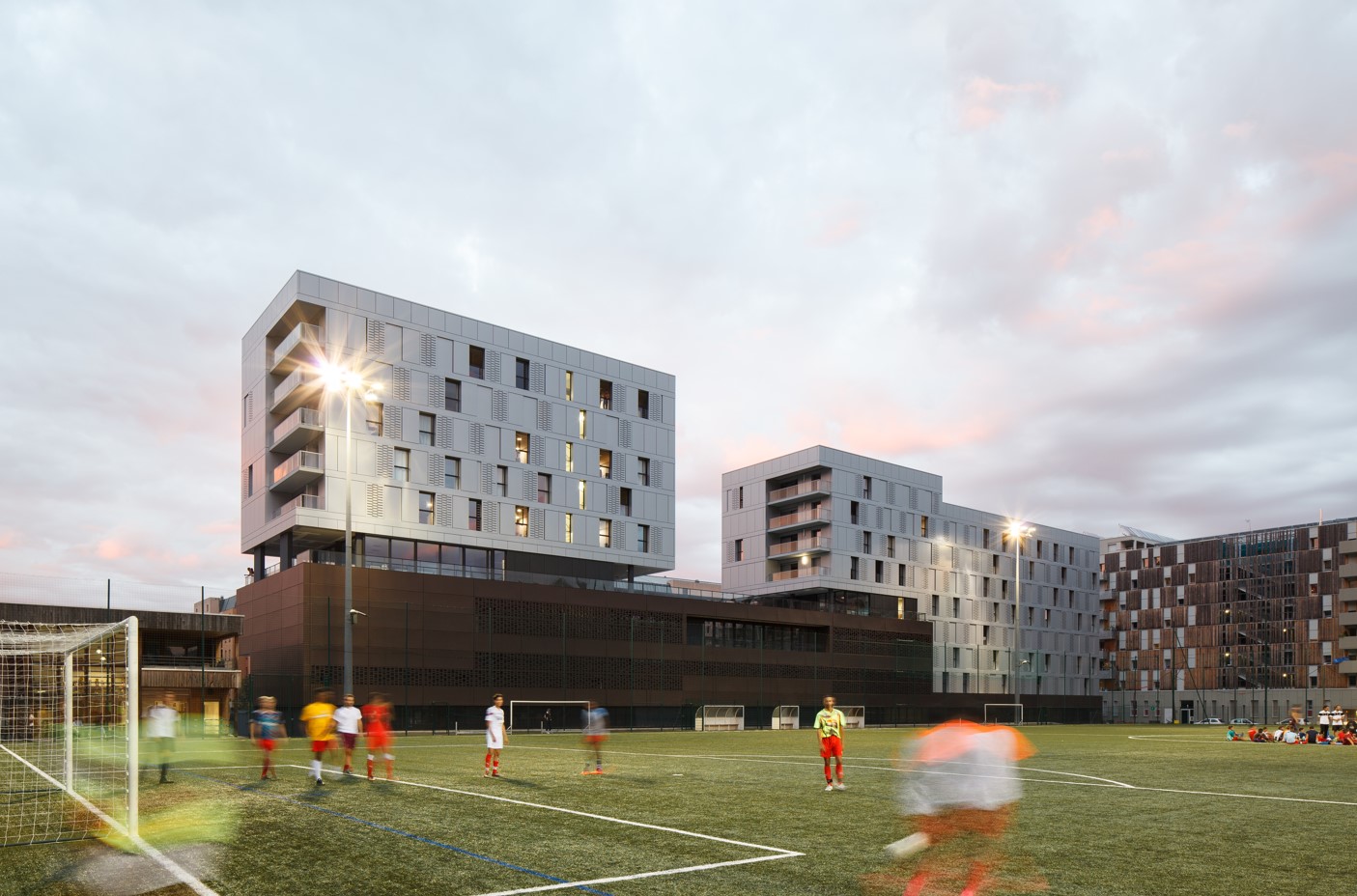 View of a football pitch with students in motion, in the evening, in front of a group of modern, well-lit buildings.
