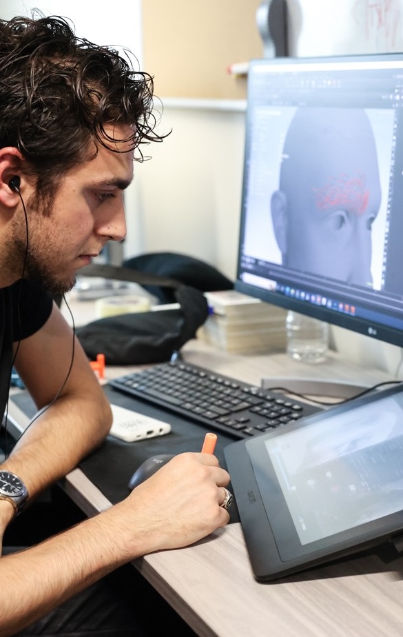 Un étudiant utilise une tablette graphique et un écran pour modéliser un visage 3D sur un logiciel spécialisé.