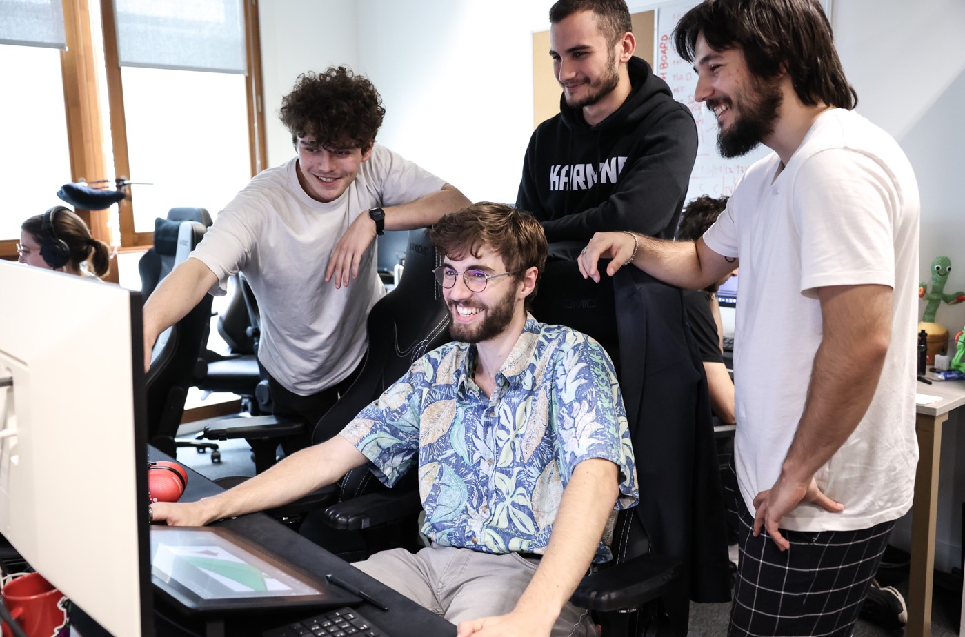Four smiling students work together at a computer workstation in a well-equipped room, concentrating on a creative project.