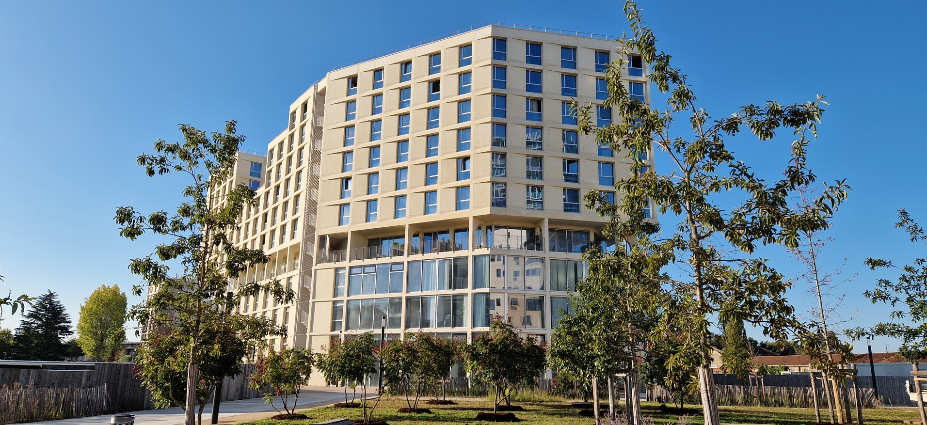 Large, modern, multi-storey building with lots of windows, surrounded by a tree-lined garden under a blue sky.